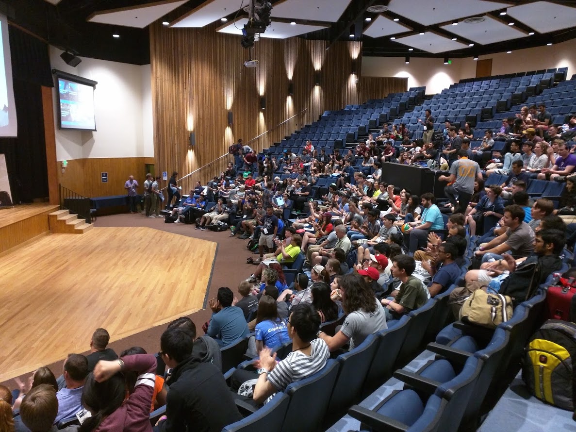 A large auditorium with around 200 people in it, with more people walking into the room to find seats.
