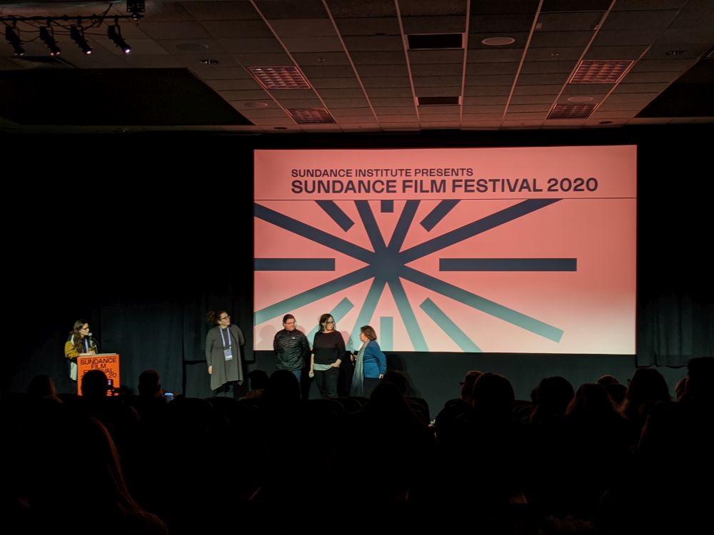 A movie theater screen, with several production members standing in front of the screen talking.
